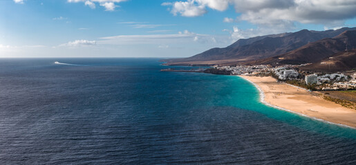 Aerial view of a curving turquoise bay, golden sand beach, and volcanic mountains on Fuerteventura near Morro Jable, with resorts, small figures, and a faint boat wake.