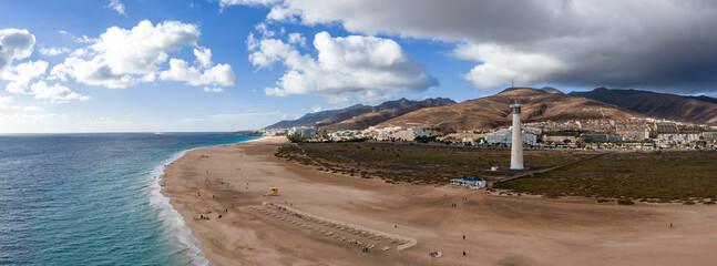 Aerial view of Playa del Matorral, Morro Jable Lighthouse, and town in Fuerteventura. Turquoise surf meets wide sands with loungers and a lifeguard post under clouds.