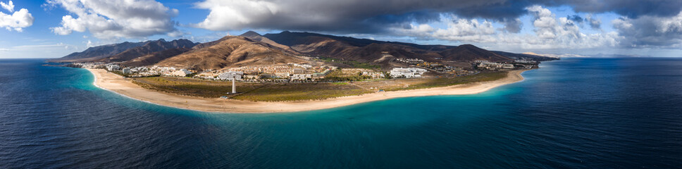 Aerial view of Playa del Matorral, Fuerteventura, with Morro Jable lighthouse. Resorts line the shore, volcanic hills rise behind, and clouds move across arid slopes. © Aerial Film Studio