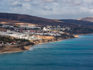 Elevated view shows whitewashed resorts and homes on dark cliffs in Morro Jable, Fuerteventura, with terraces, a church like building, and small pocket beaches. © Aerial Film Studio