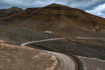 A winding dirt road curves through volcanic slopes in Fuerteventura, Canary Islands, under overcast skies, leading to a small structure as a lone cyclist rides.