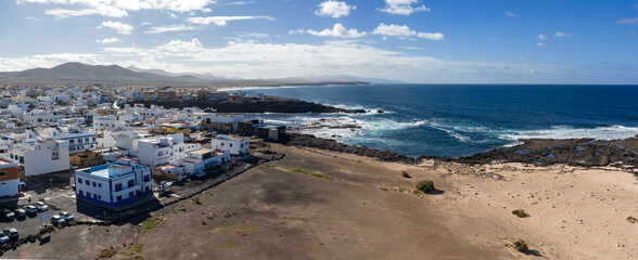 A wide aerial view shows white flat roof buildings on Fuerteventura, waves break on dark lava rock, sandy plain leads to hazy hills under bright midday light. © Aerial Film Studio