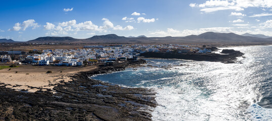 Aerial view of a whitewashed village on a rocky lava shoreline in Fuerteventura near Playa del Bajo de la Burra. Surf sparkles, arid mountains and coves frame the scene. © Aerial Film Studio