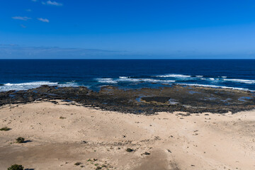Aerial midday view of Fuerteventura, Canary Islands, with pale sand, black lava reef, tide pools, and small breakers near Playa del Bajo de la Burra under a clear sky.