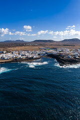 Aerial scene of whitewashed buildings by black lava rocks in Fuerteventura, Canary Islands. Deep Atlantic swells break, arid plains meet volcanic hills under midday clouds. © Aerial Film Studio