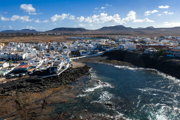 Aerial coastal town on Fuerteventura, Canary Islands, with whitewashed buildings, rocky bay, seawall, shoreline restaurants, tidal pools, and deep Atlantic waves. © Aerial Film Studio
