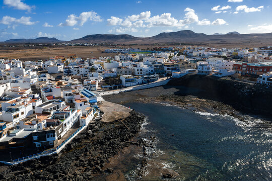 Aerial view of a coastal town on Fuerteventura, Canary Islands, near Playa del Bajo de la Burra, with white buildings, rocky harbor, tidal pools, and midday light.