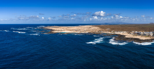 Aerial view of Fuerteventura north shore shows Atlantic surf on volcanic reefs, pale sand coves, a lighthouse near Playa del Bajo de la Burra, and distant Lanzarote. © Aerial Film Studio