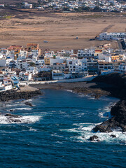 Aerial view shows whitewashed boxy buildings on lava rock coves, Atlantic waves, and arid ochre plains near suburbs in Fuerteventura, Canary Islands. © Aerial Film Studio