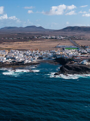 Aerial view of whitewashed buildings by a rocky inlet on Fuerteventura. Deep turquoise Atlantic waters meet arid volcanic plains, cones, and a straight roadway at midday. © Aerial Film Studio