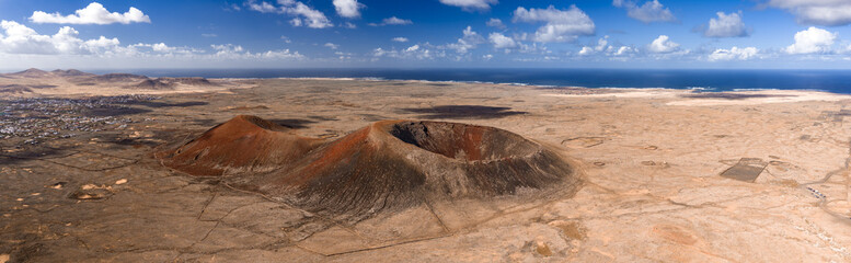 Aerial view shows volcanic cones, caldera rim, and Atlantic near Fuerteventura. Midday light reveals cinder slopes, lava paths, foot trails, and whitewashed settlements.