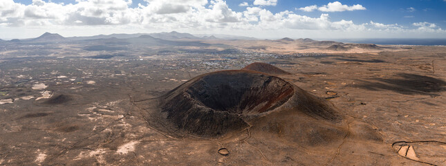Aerial view of Caldera Los Arrabales in Fuerteventura shows a dark horseshoe crater, lava fields, footpaths, a low town, ridgelines, distant cones, and the Atlantic under clouds.