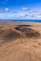 Aerial view shows a lone cinder cone on arid plains in Fuerteventura, Canary Islands. Atlantic Ocean shimmers, lava fields and faint hiking tracks encircle the base.