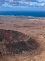 Aerial view shows a rust colored cinder cone near Fuerteventura, Canary Islands, with steep rim, rubble paths, flat lava fields, Atlantic horizon, and a small coastal settlement.