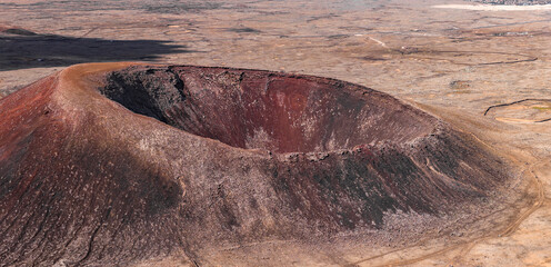 Aerial view shows a near perfect crater in Fuerteventura, Canary Islands. The rim has rust red and charcoal black rocks, steep caldera layers, and faint hiking tracks.