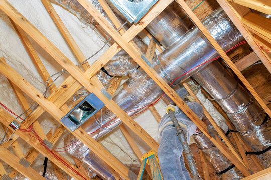 Workers install ventilation ducts between wooden beams in home under construction, utilizing tools ladders.