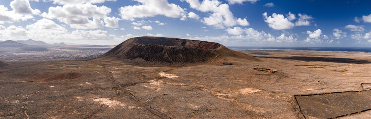 A wide aerial view shows Caldera Los Arrabales in Fuerteventura, a dark rimmed cone, lava fields, a town by low mountains, and the Atlantic under midday cumulus.