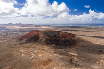 Aerial view of a volcanic caldera with rust red and charcoal slopes on Fuerteventura, low white towns, distant dunes, and the Atlantic coast under scattered cumulus clouds.
