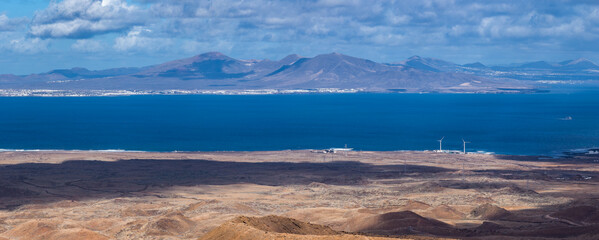 Aerial scene shows ochre hills, lava plains, two wind turbines, a coastal facility, and a lone boat near Fuerteventura, facing Lanzarote towns under midday light.