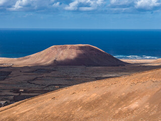 A rounded volcanic cone rises from an ochre plain in Fuerteventura as the deep Atlantic stretches behind, waves trace the distant shore, warm light and clouds reveal smooth flanks.