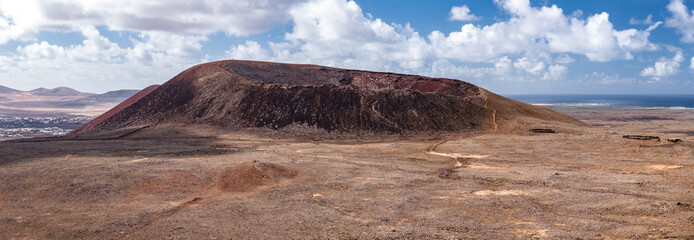 Rust red cone with horseshoe crater rim rises from lava plain, Atlantic gleams beyond. Small settlements sit at left, tiny hikers trace footpaths in daylight tones.