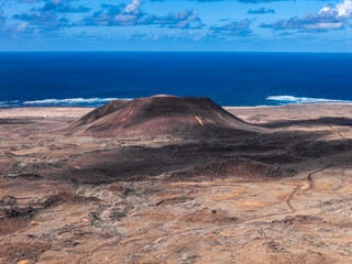Aerial view shows a solitary volcanic cone and flat topped crater on Fuerteventura, Canary Islands. Ash streaked slopes meet ochre lava plain and cobalt Atlantic at midday.