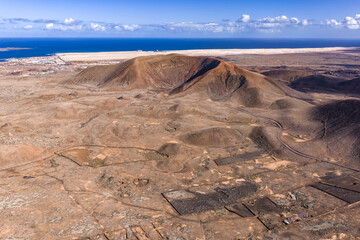 Aerial view shows a reddish cinder cone, smaller cones, and lava fields near Corralejo, Fuerteventura. Pale dunes meet the deep Atlantic, with a winding road in bright midday light.