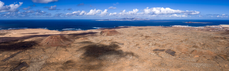 Arid volcanic plains with rust cinder cones stretch in northern Fuerteventura, Canary Islands. Atlantic horizon shows Lanzarote and Lobos near Corralejo at midday.