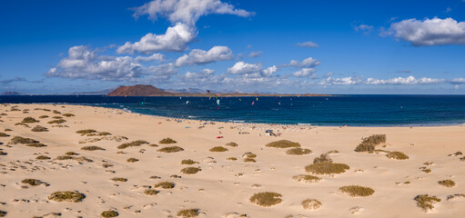 Aerial view of Corralejo dunes and Atlantic in Fuerteventura. Kite surfers ride, sunbathers rest on the shore. Isla de Lobos and Lanzarote rise under crisp daylight.