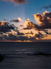 Sunbeams fan through broken clouds over the Atlantic near Fuerteventura. Rugged shoreline and open horizon frame dark rippled water, glowing cloud edges, and strong contrast.