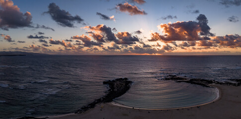 Aerial view shows crescent lagoon and lava rock jetty at Playa del Bajo de la Burra, Fuerteventura, Atlantic swells, sandy beach, orange pink horizon, rays, Lanzarote silhouettes.