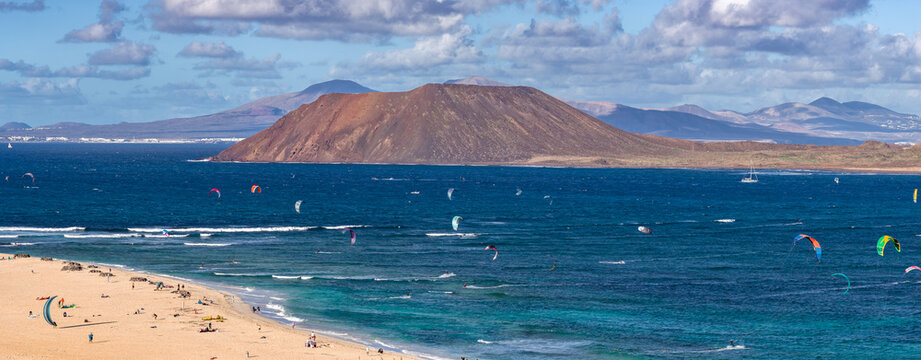Kitesurfers skim turquoise waters by Corralejo near Playa del Bajo de la Burra in Fuerteventura, with Isla de Lobos and Lanzarote mountains under crisp daylight skies.