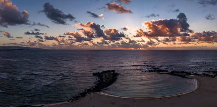 Aerial view shows crescent lagoon and lava rock jetty at Playa del Bajo de la Burra, Fuerteventura, Atlantic swells, sandy beach, orange pink horizon, rays, Lanzarote silhouettes.