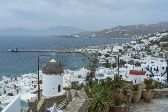 View of whitewashed buildings cascade down the hills towards the turquoise sea, a classic windmill standing sentinel against the cloudy sky, Mykonos, Greece.