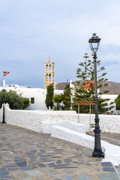 View of a classic black iron street lamp stands tall against the backdrop of a white building with a tower, under a soft sky, Ano Mera, Mykonos, Greece.
