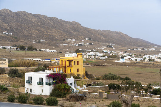 View of a yellow building stands out against the backdrop of a rocky hill and scattered white houses in a dry, sun-soaked landscape, Ano Mera, Mykonos, Greece.