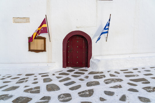 View of a vivid red door contrasts against the stark white walls and patterned cobblestone street, Monastery of Tourliani, Mykonos, Greece.