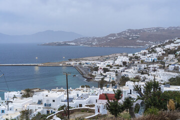 View of a coastal town with white buildings meeting the sea, a pier extending into the water under a cloudy sky, Mykonos, Greece.