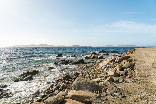 View of turquoise waves crash against the rocky shore, meeting a concrete path under a pale blue sky, a serene coastal scene, Mykonos, Greece.