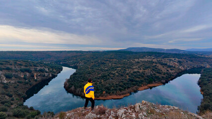 Adventurous hiker in a yellow jacket standing on the edge of a cliff, contemplating the stunning view of the Duero River canyon from the Mirador Curvas del Duero in Soria, Castilla y León, Spain.
