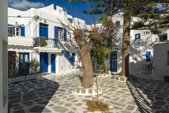 View of sun-kissed white buildings with striking blue doors and window frames, and a patterned stone courtyard, Old Town, Mykonos, Greece.