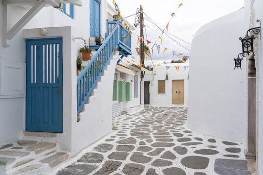 View of the rhythmic pattern of cobblestone streets winding through whitewashed buildings with vibrant blue accents under a sky adorned with festive flags, Old town, Mykonos, Greece.