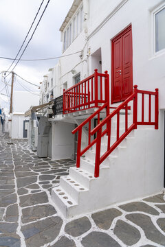 View of vibrant red accents pop against stark white buildings and cobblestone streets under a cloudy sky, creating a striking contrast, Old town, Mykonos, Greece.