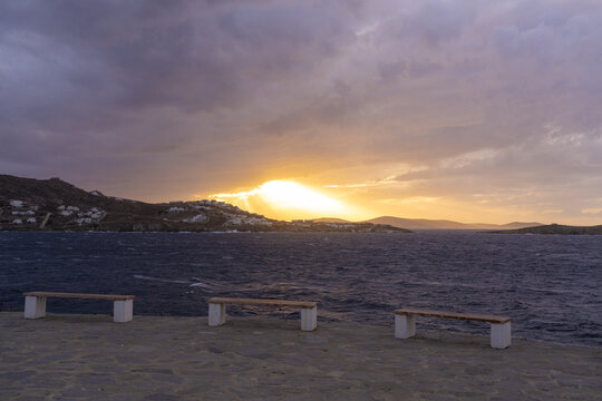View of the sun breaking through storm clouds over the tranquil sea, casting a golden glow on the distant hills, as seen from the empty benches, Mykonos, Greece.