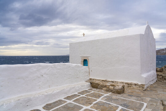 View of a whitewashed chapel with a simple cross gleams against the turbulent sea under a stormy sky, a classic Cycladic scene, Mykonos, Greece.