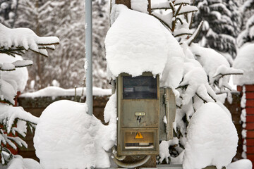 An outdoor electrical meter covered with thick snow stands on a pole in a winter landscape. Snow-laden spruce branches and a brick fence surround the scene, emphasizing cold weather and rural life