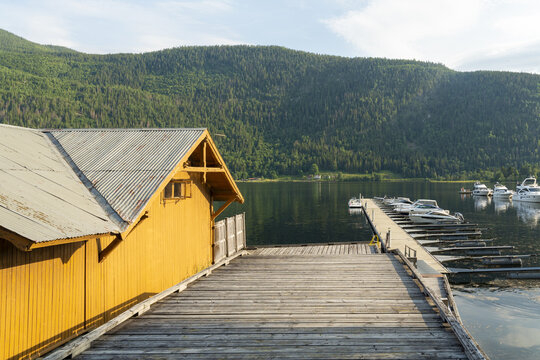 View of a yellow boathouse extends onto a wooden pier leading to a serene lake dotted with boats, against a backdrop of verdant mountains, Dalen, Telemark, Norway.