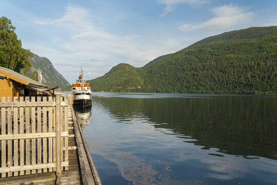 View of a vintage steamboat docked at a wooden pier, reflecting calmly on the water, surrounded by verdant, forested mountains, Dalen, Telemark, Norway.