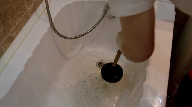 Individual using a black plunger to clear a clogged bathtub drain, showcasing the action of plunging and the gradual removal of debris from the tub surface