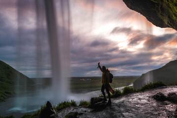 View of a person in yellow jacket raising their hand in front of the powerful Seljalandsfoss waterfall against a dramatic sunset sky, South Island, Iceland.